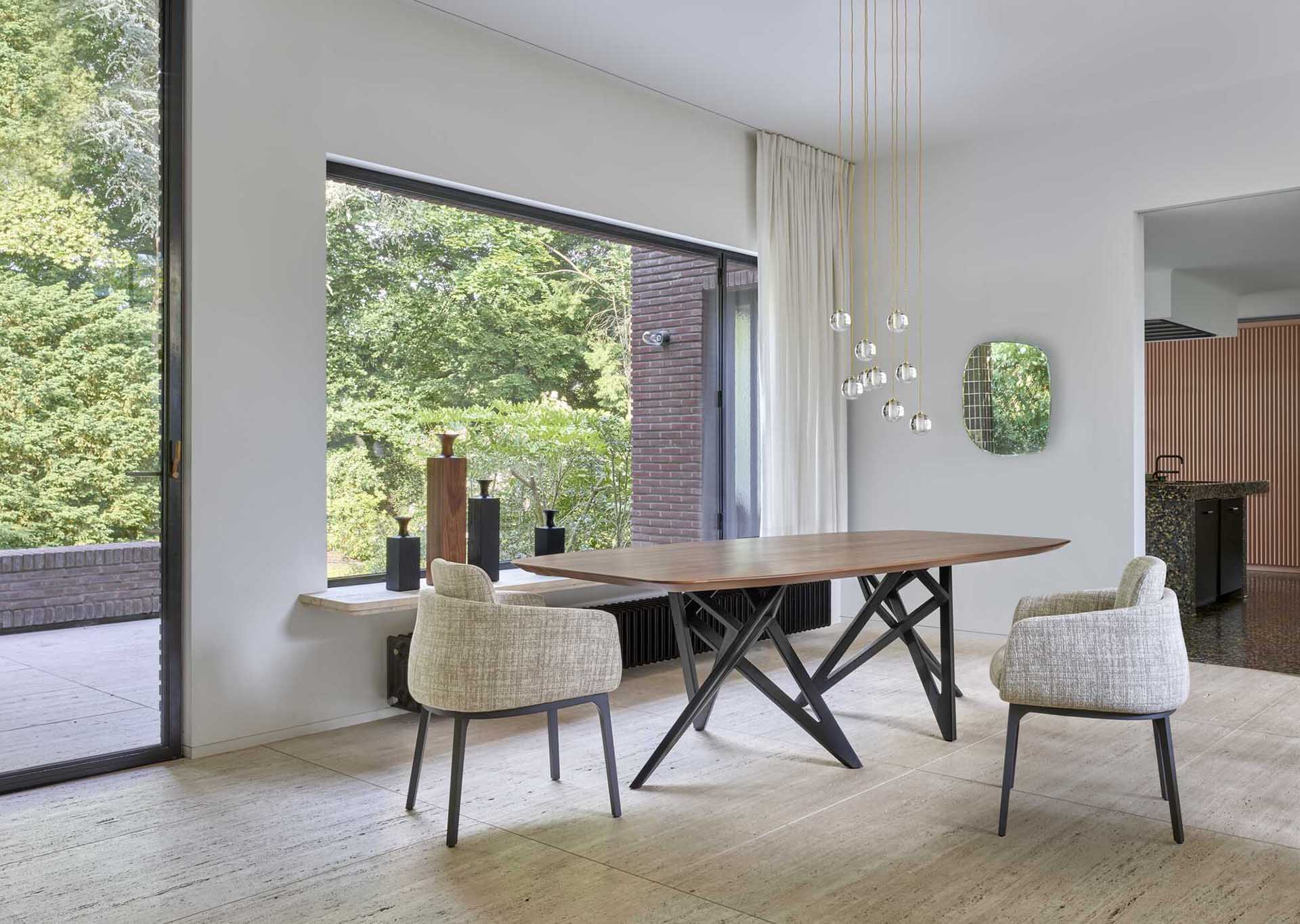 Modern dining area with rectangular wooden table, two upholstered chairs, hanging glass pendant lights, and large window overlooking green trees.