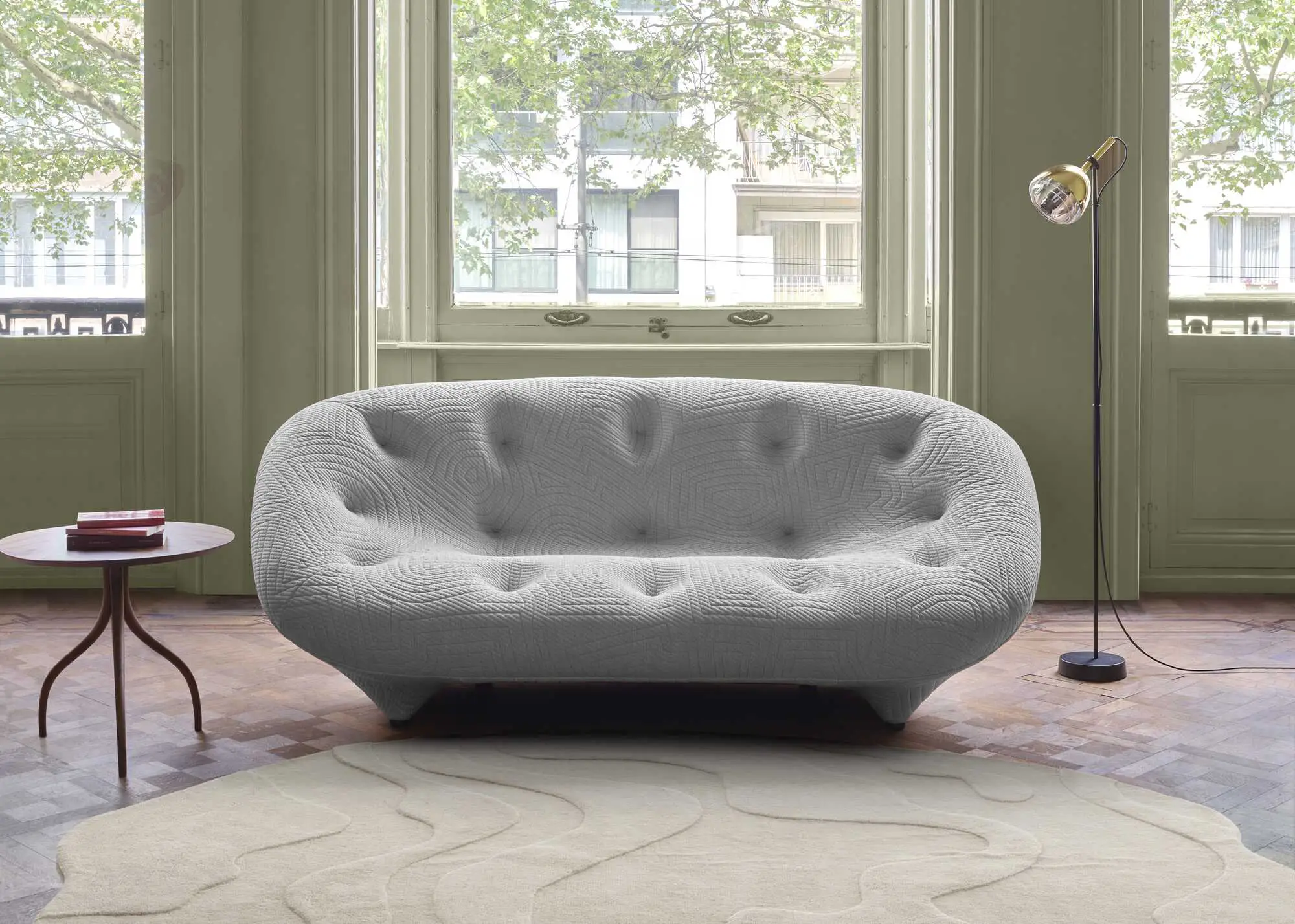 Modern grey quilted sofa with tufted design in front of large windows, accompanied by a round wooden side table with books and a tall black floor lamp with a gold lampshade.