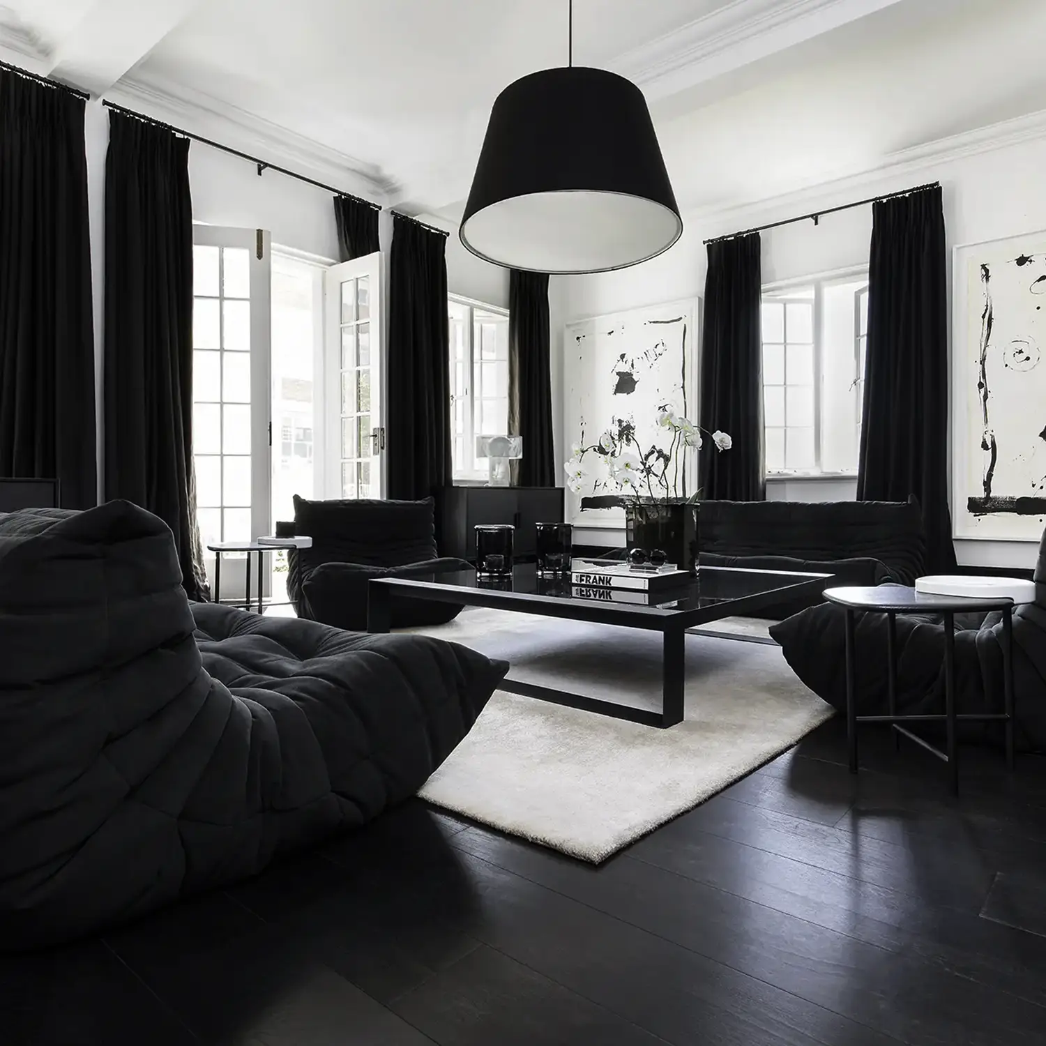Modern living room with black cushioned lounge chairs, black curtains, a black coffee table on a white rug, and large windows with white frames.